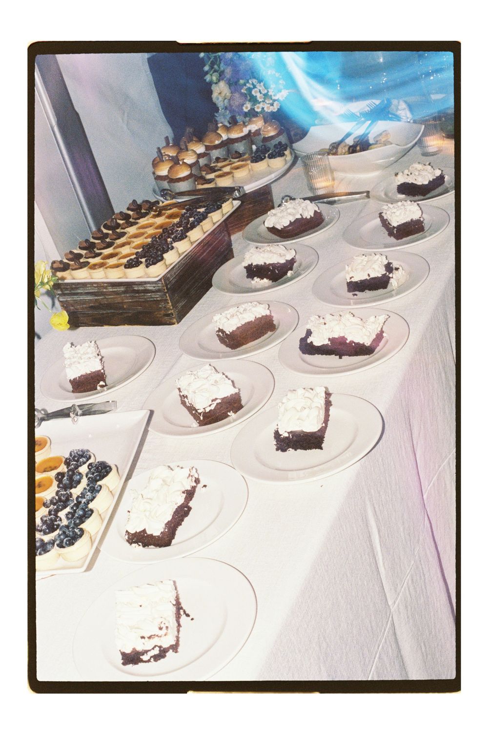 A dessert table with chocolate cake slices topped with white frosting on clear plates, alongside blueberry pastries at a catered event.