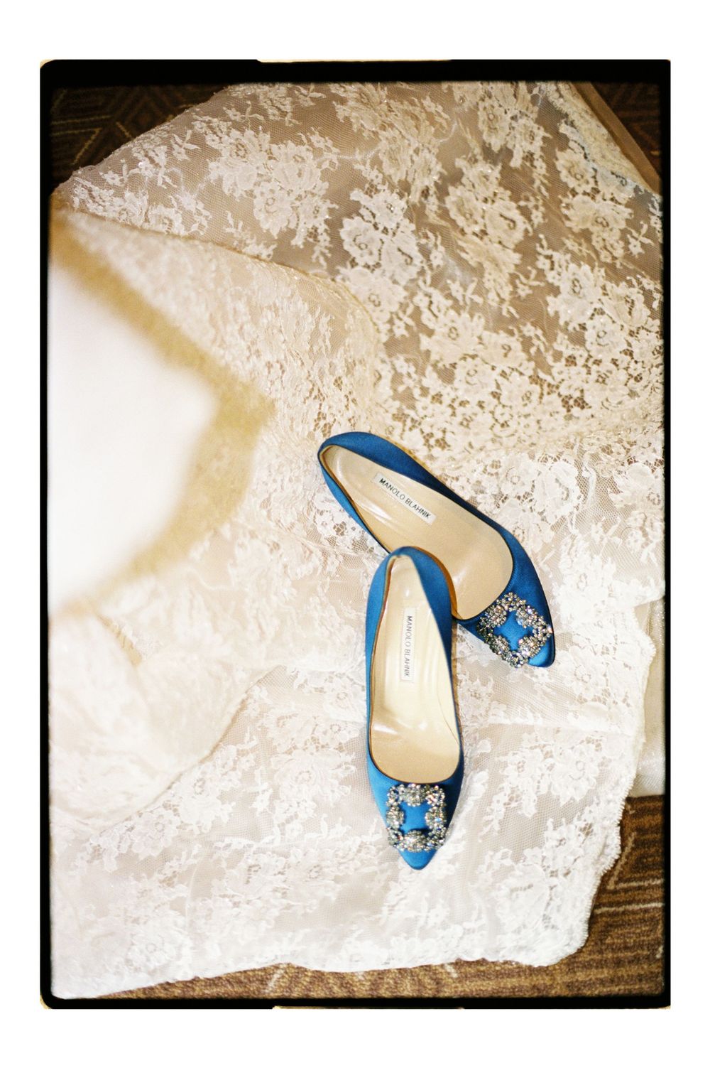 Blue satin Manolo Blahnik heels with jeweled buckles resting on a white lace wedding dress.