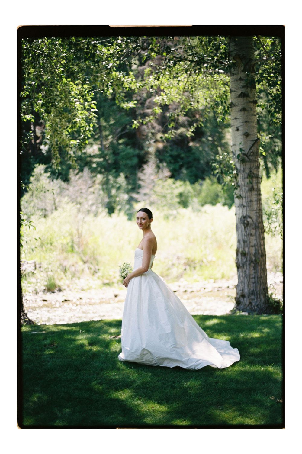 Bride in white strapless ball gown holding bouquet standing among green trees on lush grass on sunny day.
