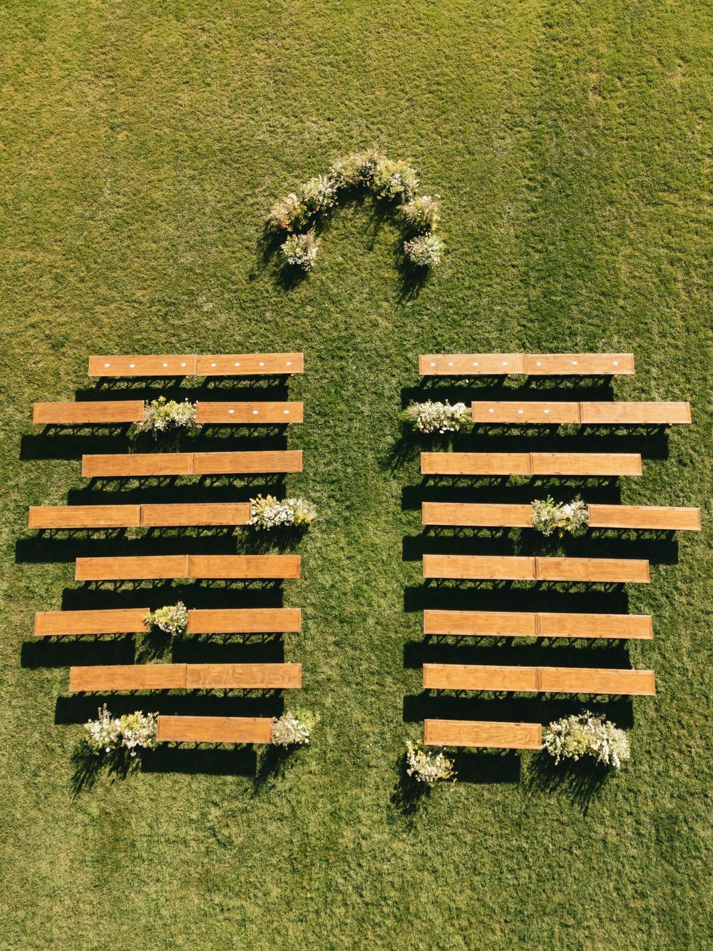 Aerial view of wooden benches arranged in rows on green grass with floral decorations for an outdoor wedding ceremony.