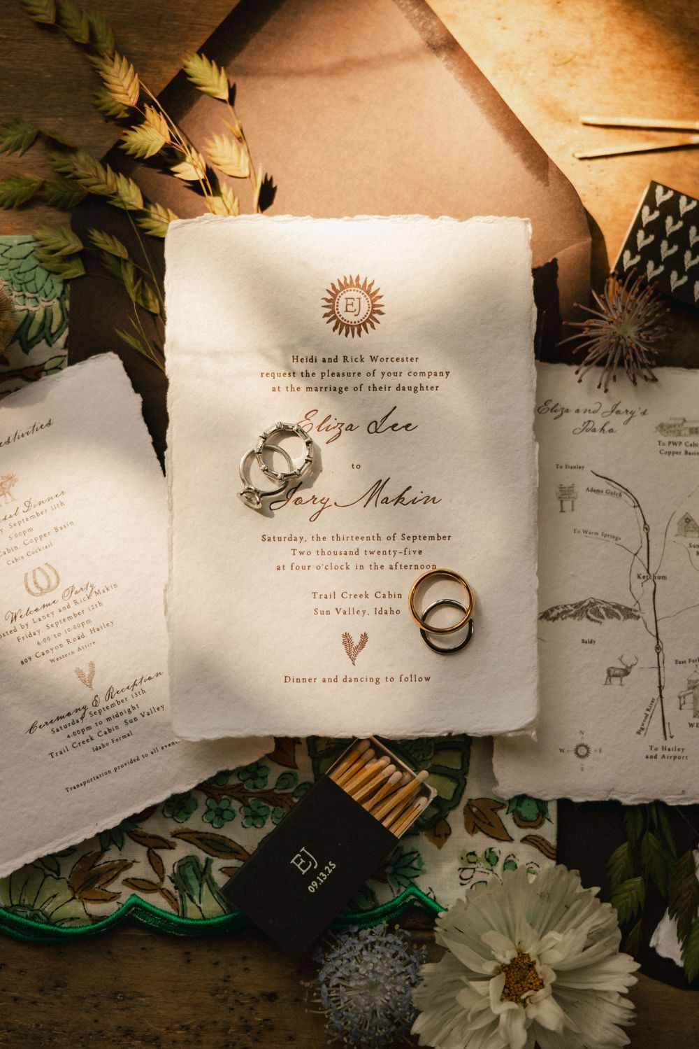Elegant wedding invitation suite with gold calligraphy, wedding rings, and floral accents on a rustic wooden surface.