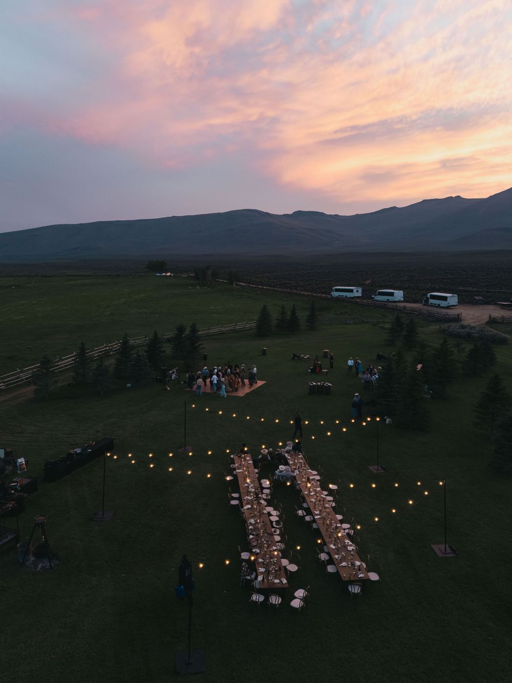 Aerial view of an outdoor wedding reception with string lights at sunset, set in a scenic mountain meadow.