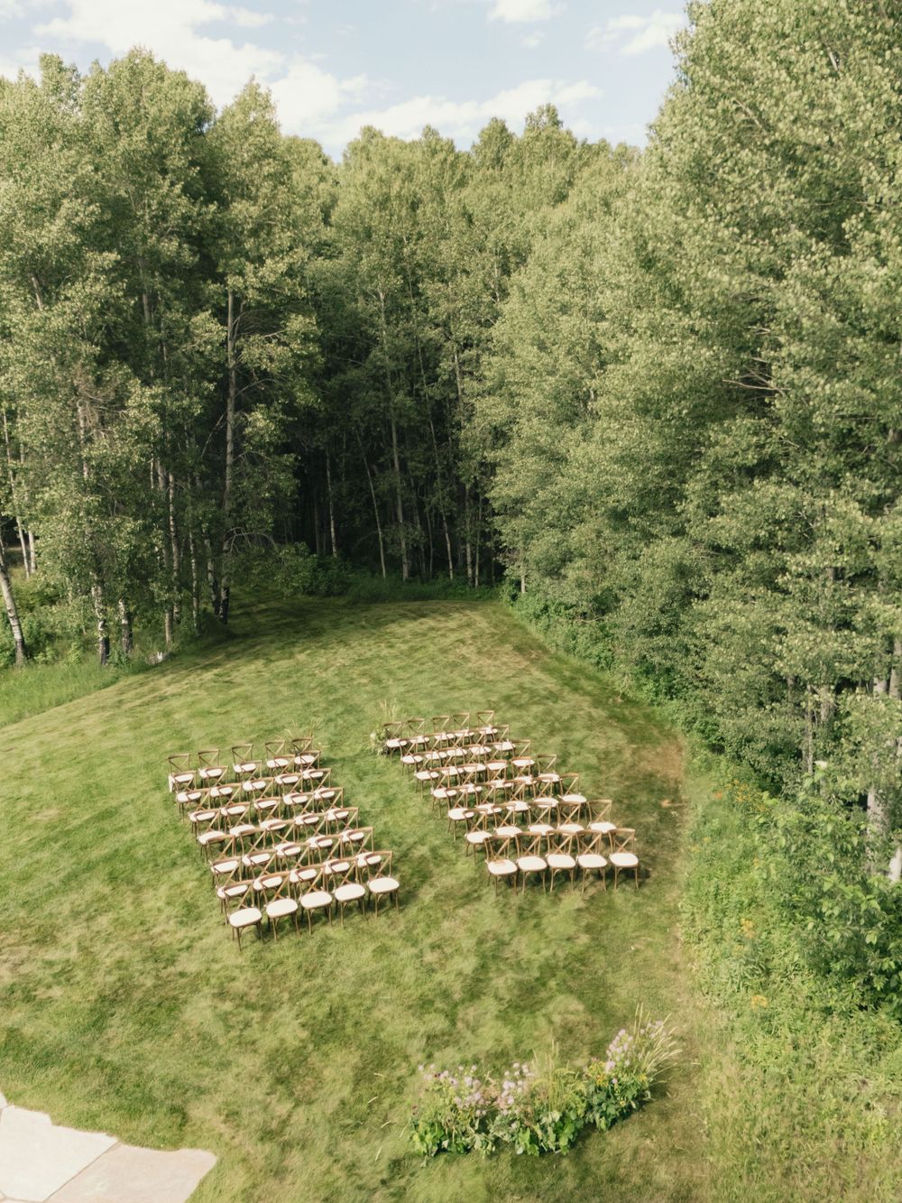 Aerial view of outdoor wedding ceremony chairs arranged in rows on a green lawn surrounded by tall aspen trees.