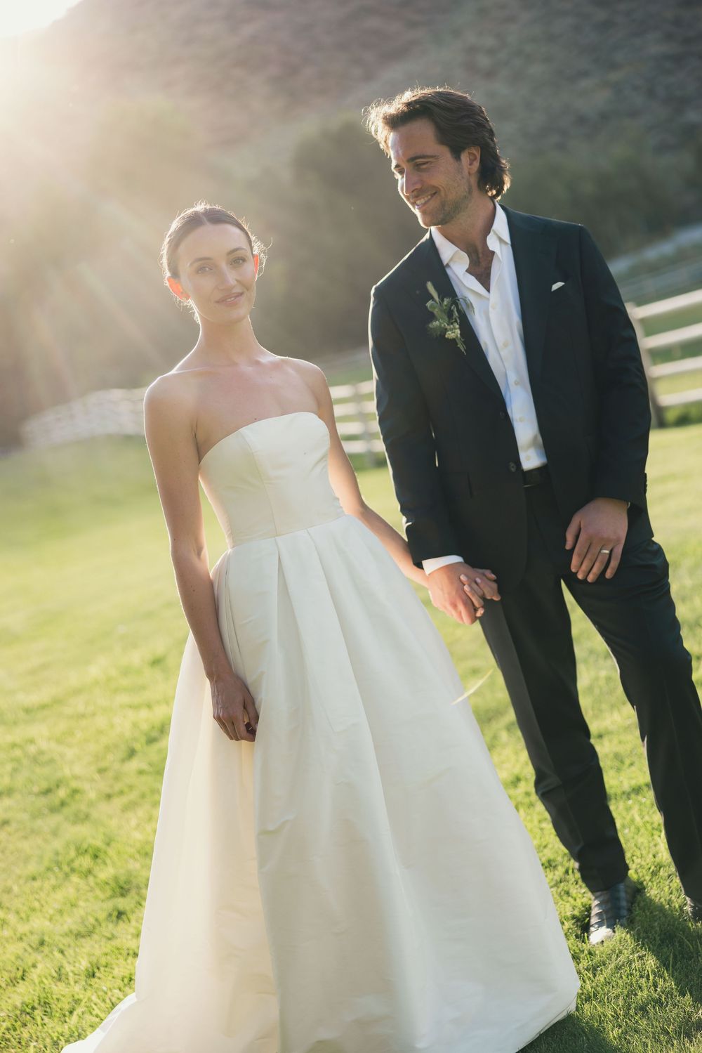 A bride in a strapless white gown and groom in a dark suit hold hands outdoors on green grass in golden sunlight.