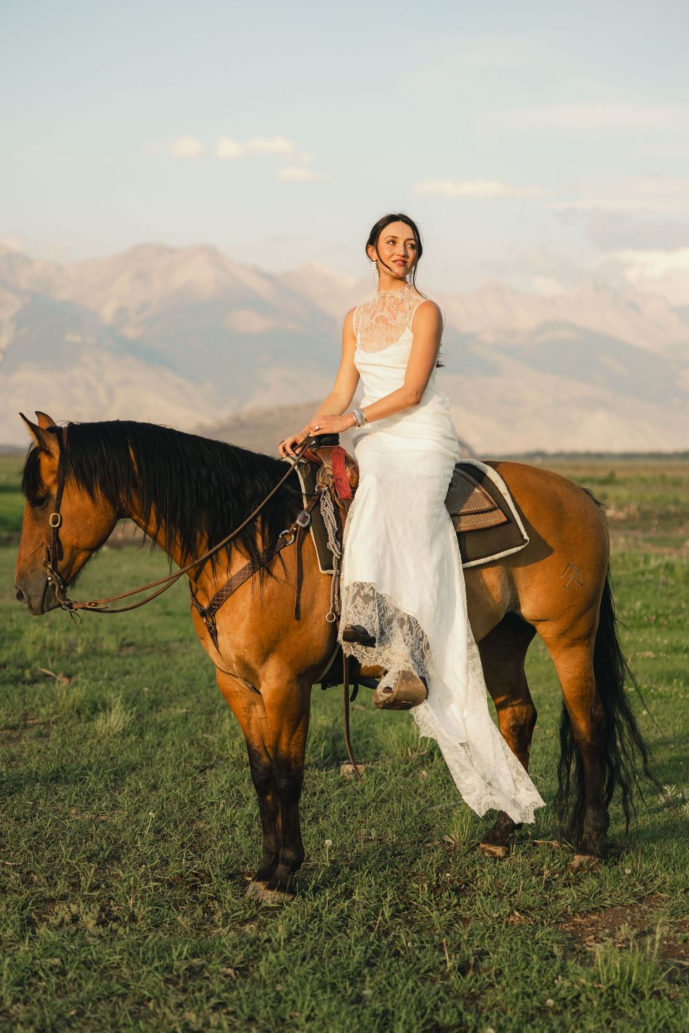 Bride in white gown riding a brown horse in a green field with mountains in the background.
