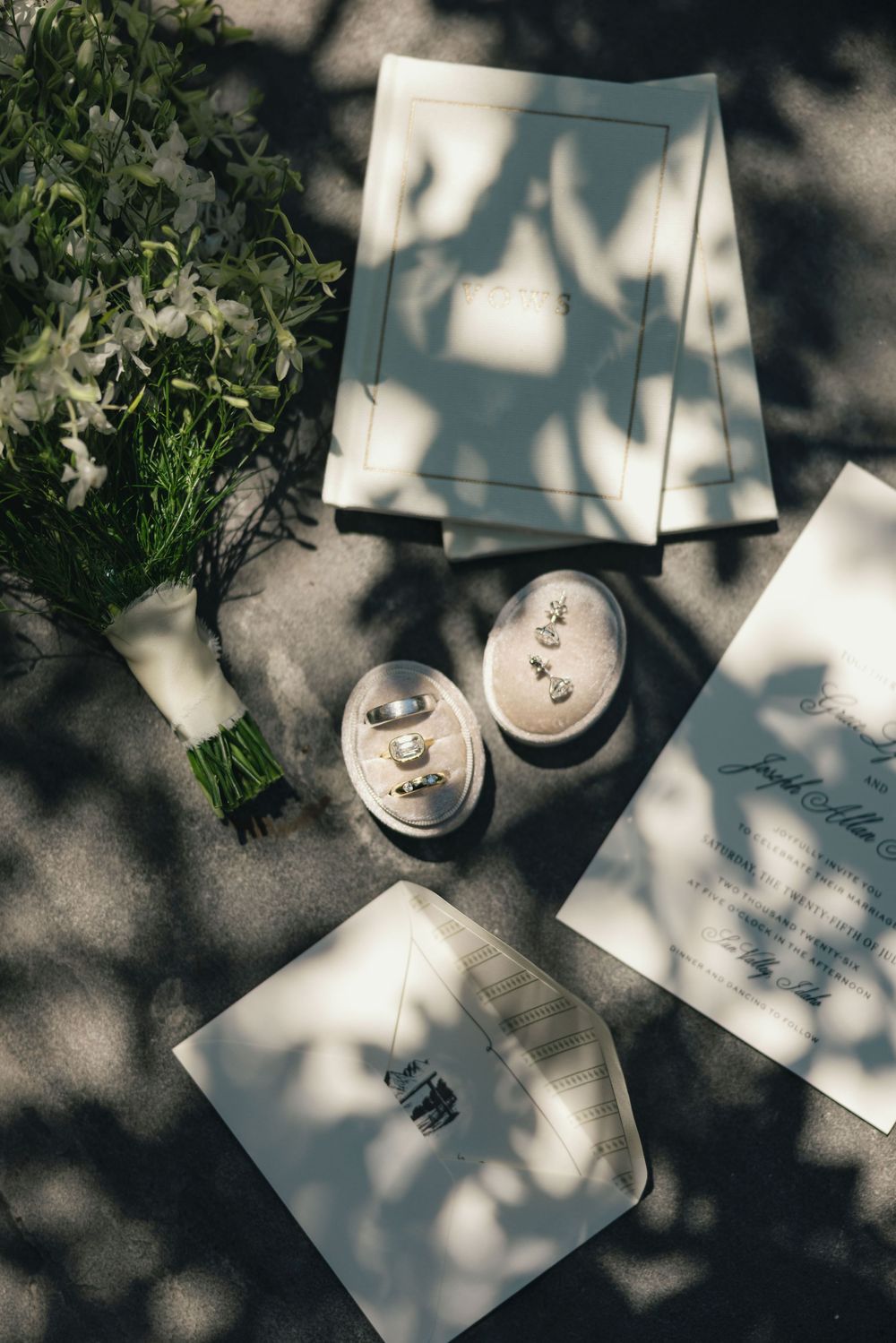 Wedding flat lay with rings, bouquet, invitation cards, and dappled sunlight shadows on gray surface.