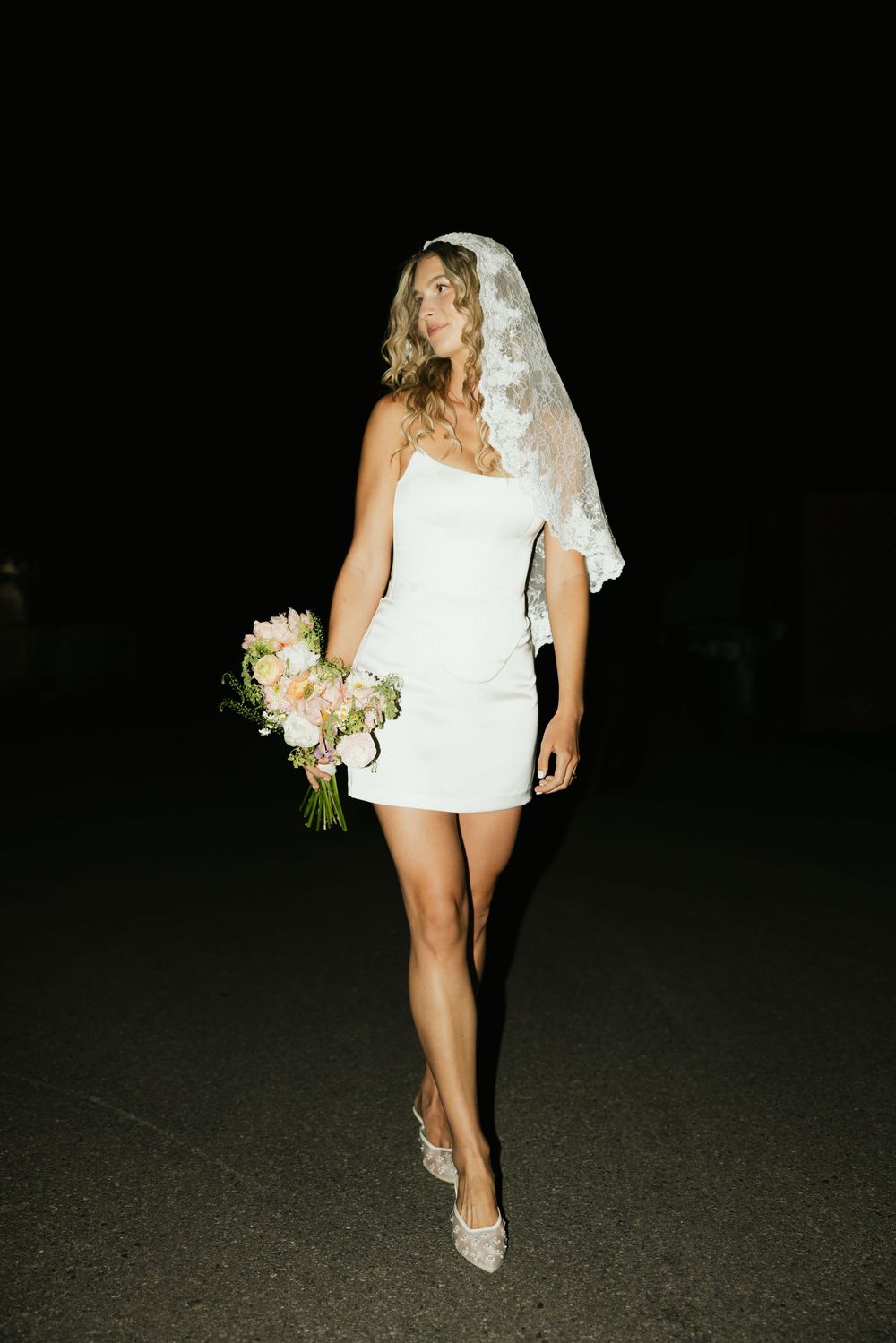 Bride in short white dress and veil walking with bouquet at night.