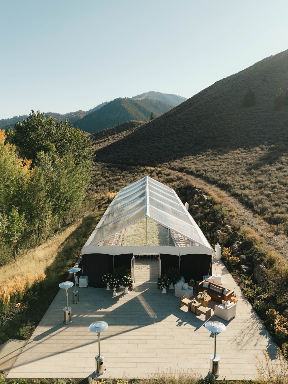 Aerial view of a clear tent venue on a wooden deck surrounded by mountains and trees under a bright blue sky.
