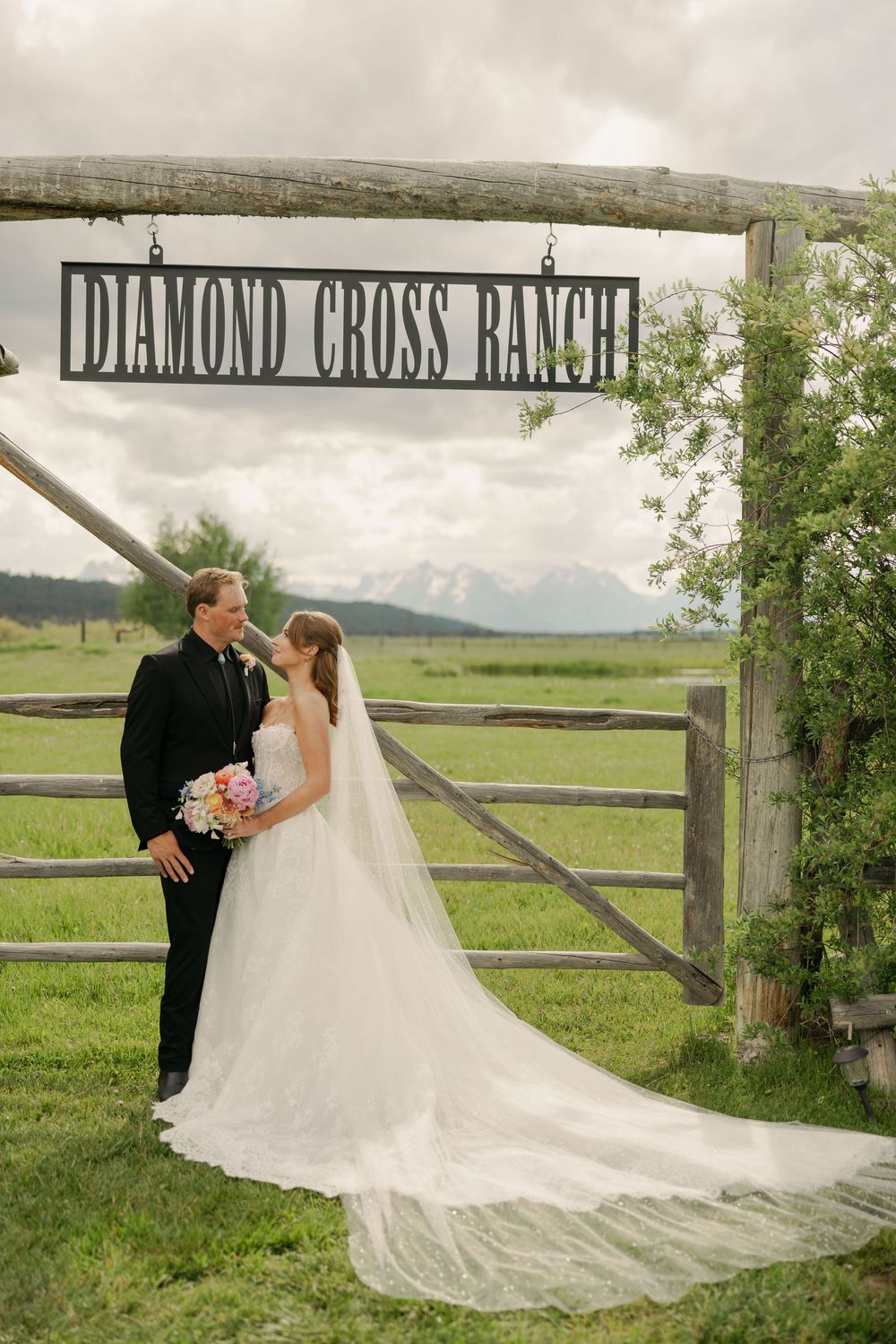 Bride and groom pose under Diamond Cross Ranch wooden sign gate on lush green Wyoming ranch wedding day.