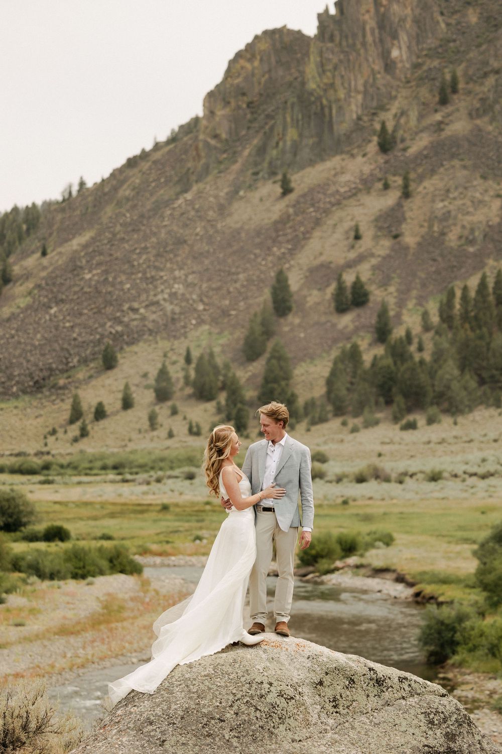 Couple sitting on a large rock in wedding attire with dramatic mountain landscape in the background.
