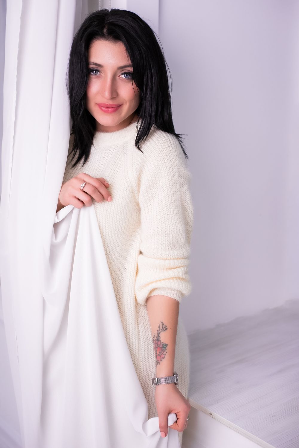 Woman in cozy white sweater poses against light background showing arm tattoo in casual photoshoot series.