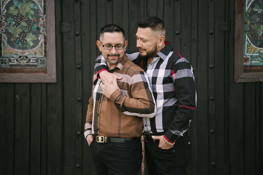 Two stylishly dressed men in vintage-inspired outfits pose together against a dark wooden wall with decorative panels.