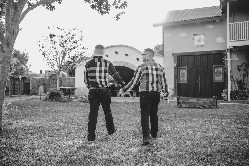 Two people in matching plaid shirts hold hands while walking on a grassy lawn with a vintage camper in the background.