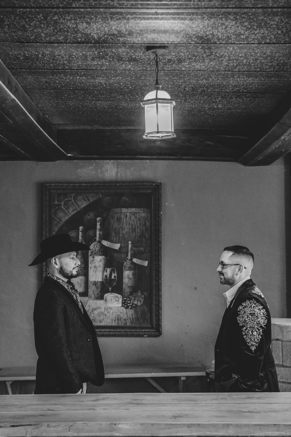 Two people in contrasting outfits stand facing each other in a dimly lit room with a hanging lantern and framed artwork.
