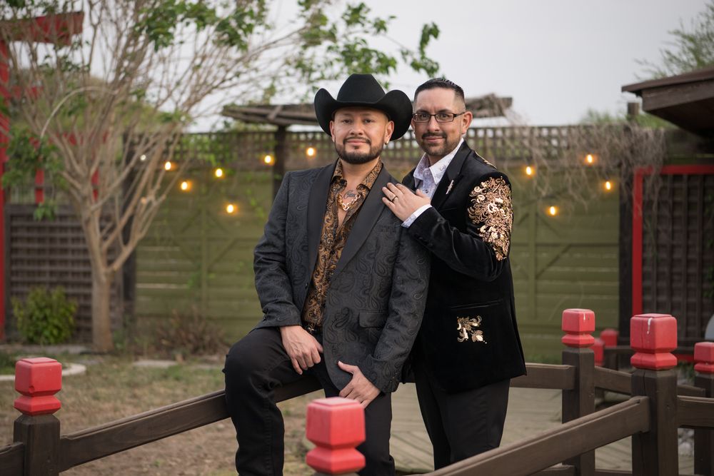 Two people in formal attire pose together outdoors at dusk with string lights and red decorations visible.