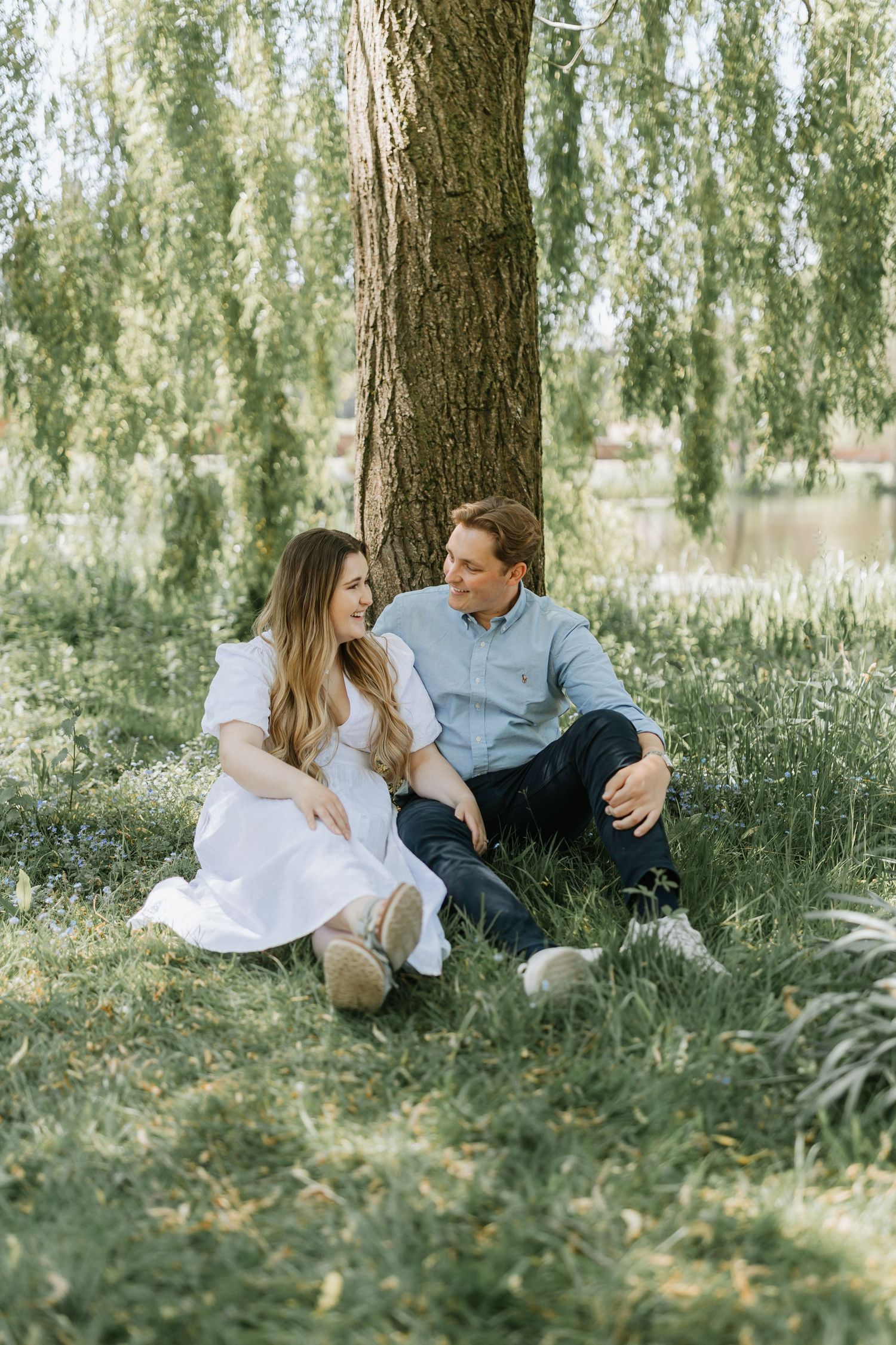 A couple relaxes together on green grass beneath willow trees in a romantic outdoor setting.