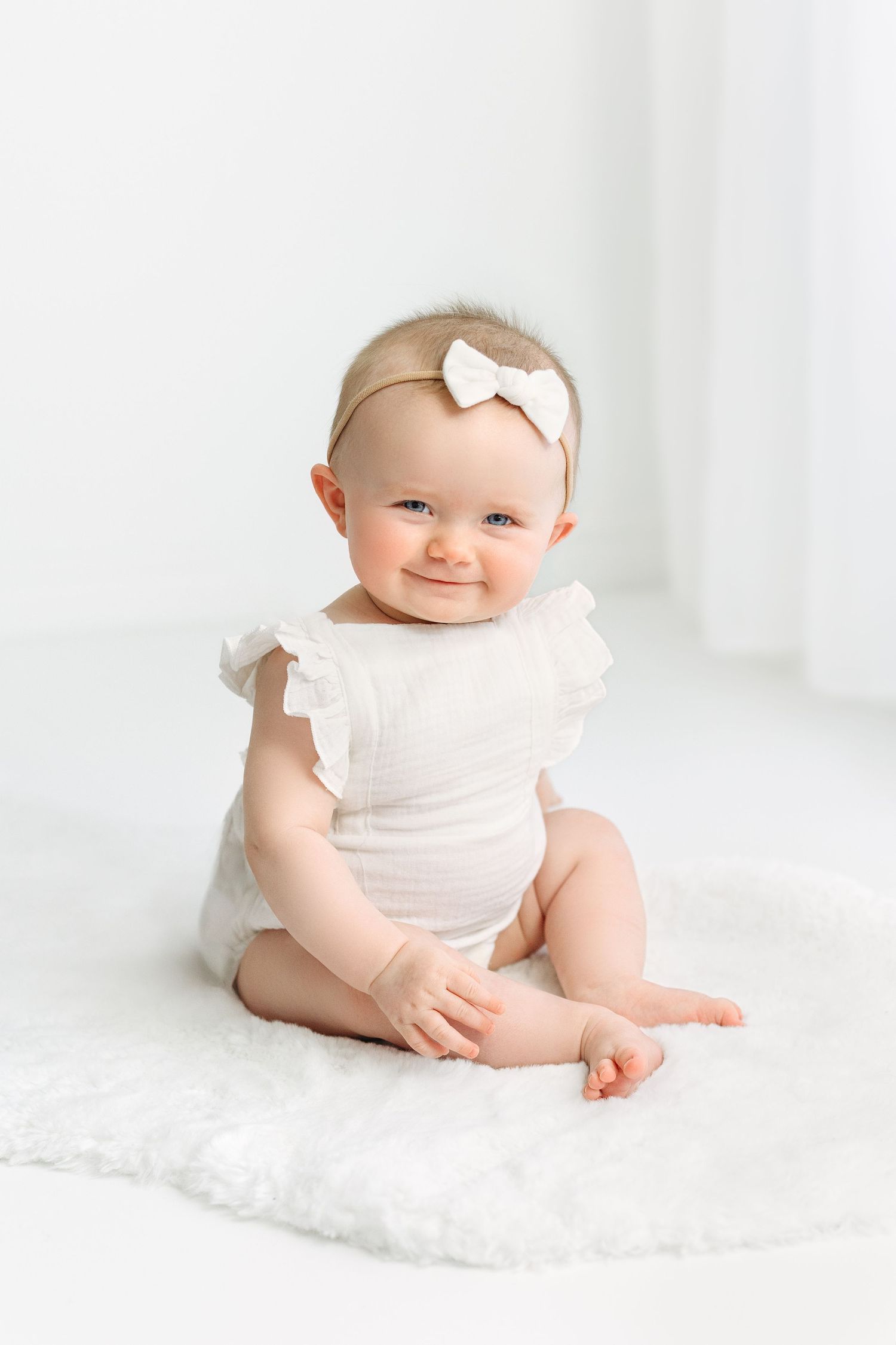 Cheeky smile on a 6 month old baby girl with white frilly onesie and bow. Sitting on a white fur rug.