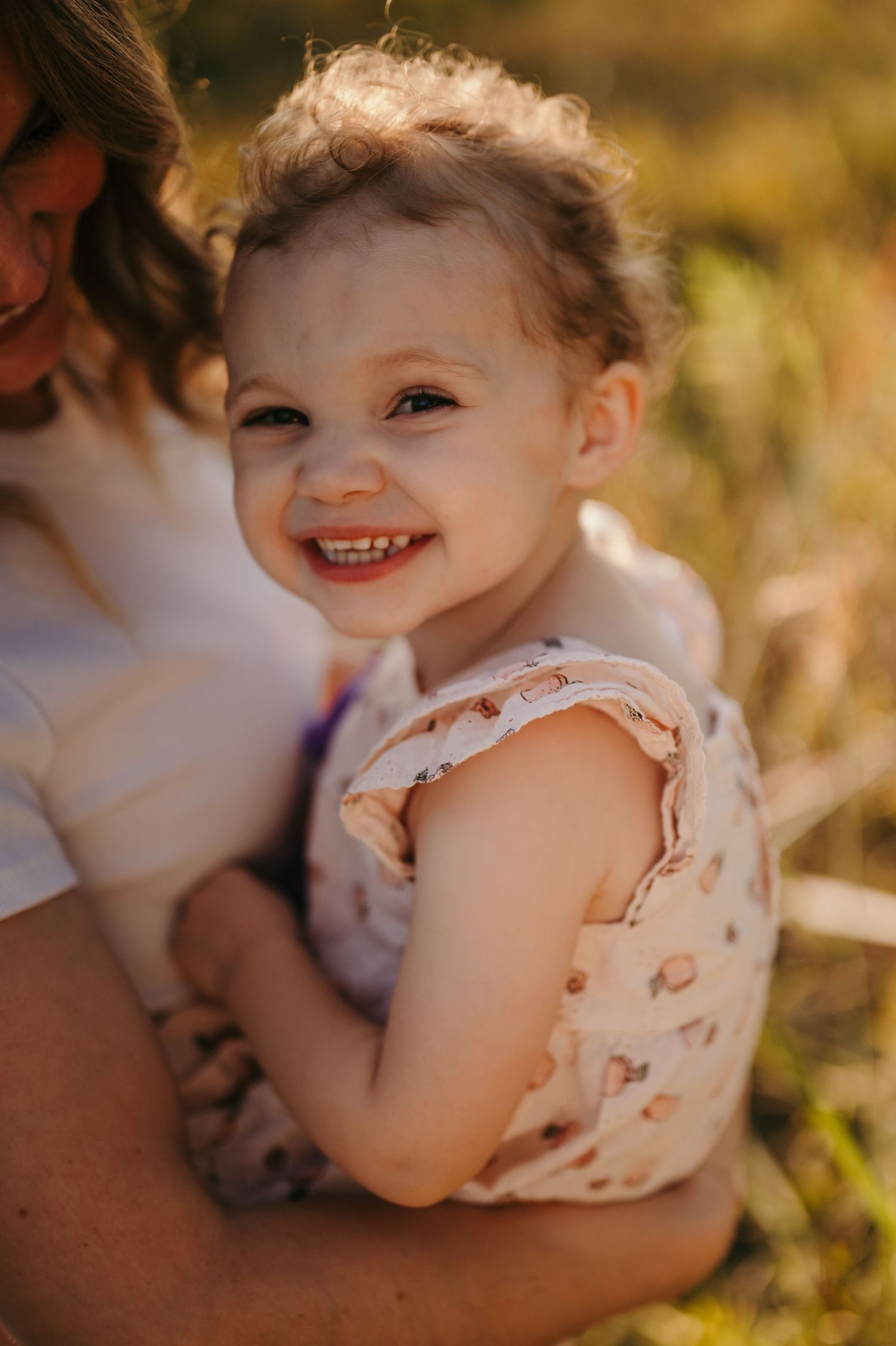 Toddler girl smiling up at the camera in golden hour light by tall grass
