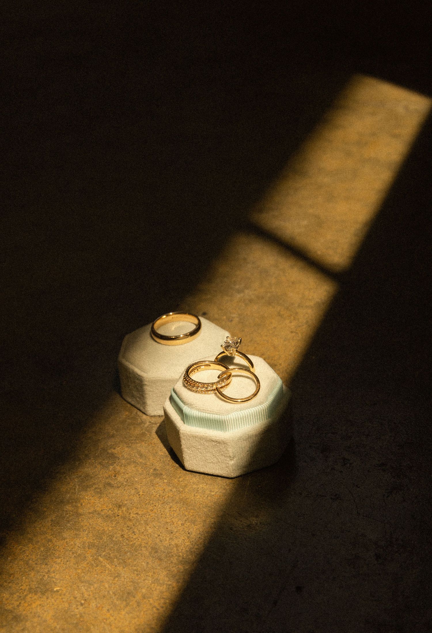Two gold wedding rings sit in white hexagonal ring boxes illuminated by dramatic sunlight.