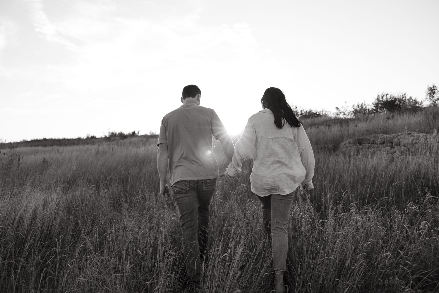A couple walks hand in hand through a sunlit field in black and white.