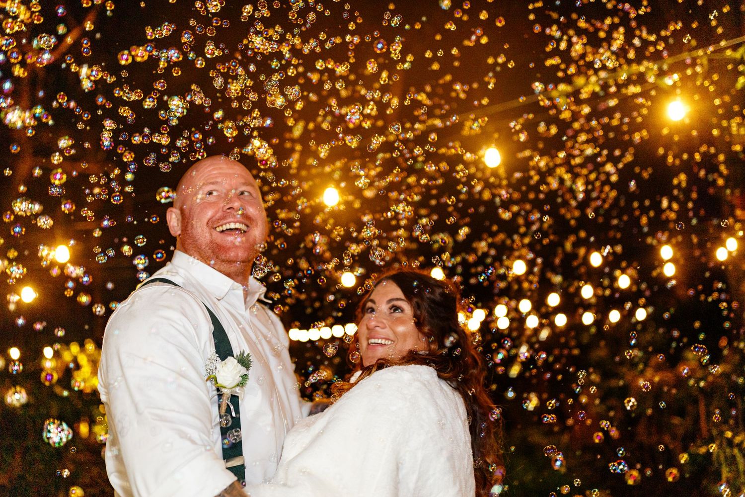 A couple laughs joyfully under twinkling string lights and bubbles creating a magical nighttime  atmosphere. The Love Shack Knutsford.