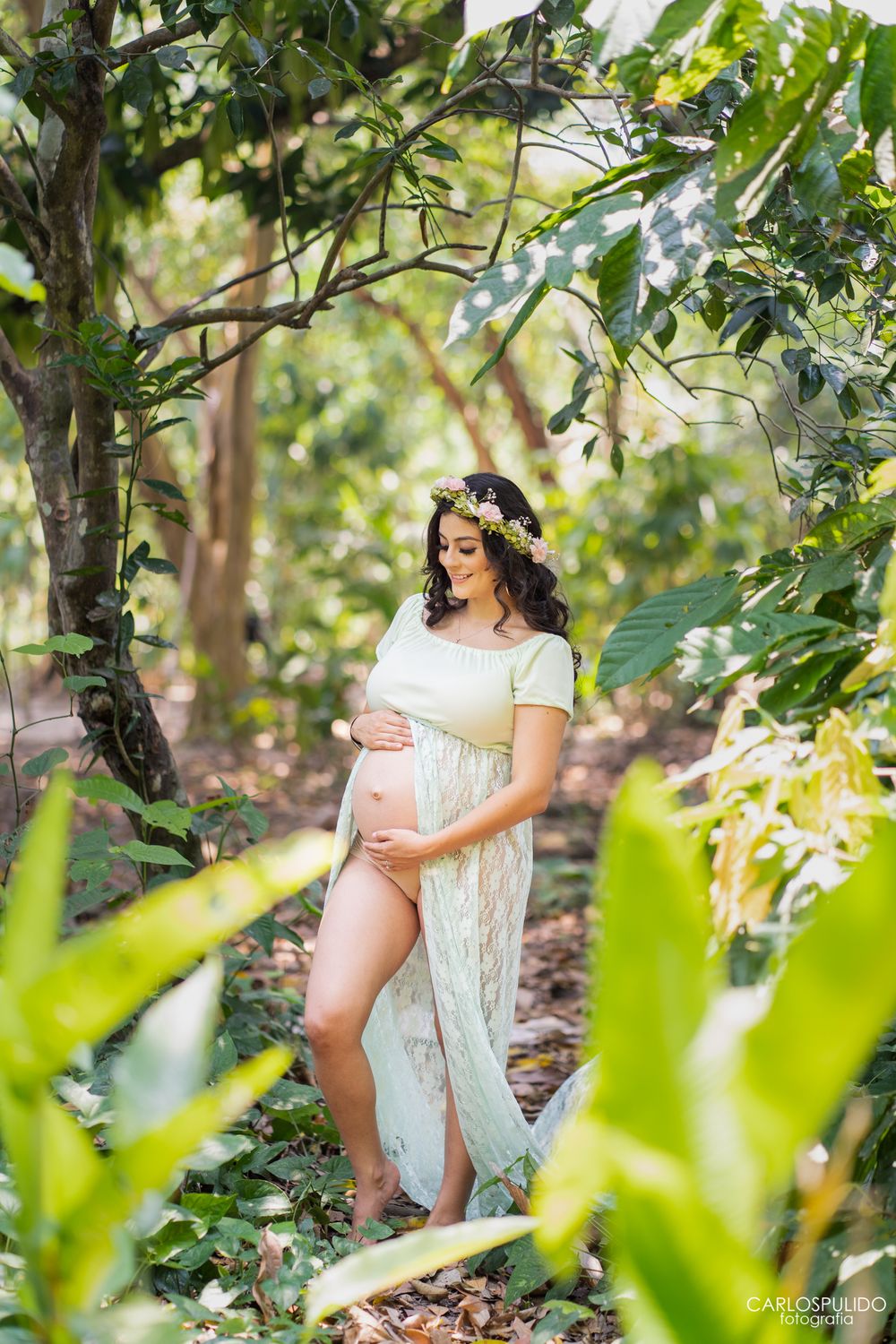 Maternity photo shoot in a lush garden setting with sunlit foliage and a flowing white lace dress.