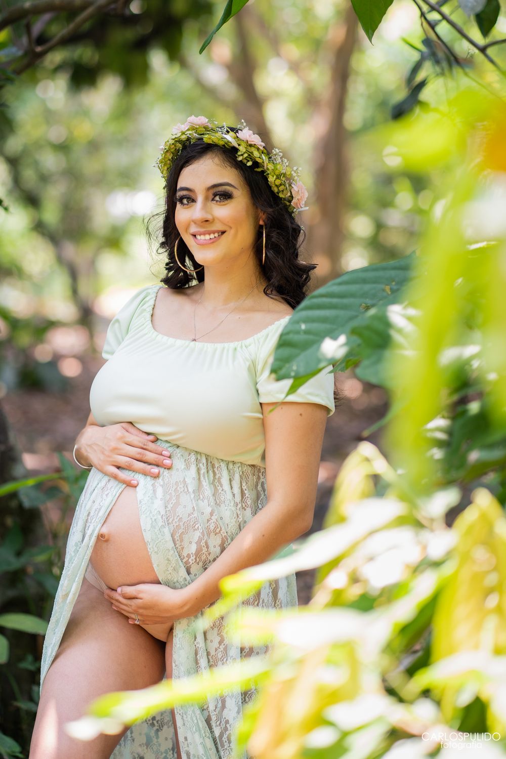 Expecting mother wearing a floral crown and white dress poses elegantly among tropical greenery.