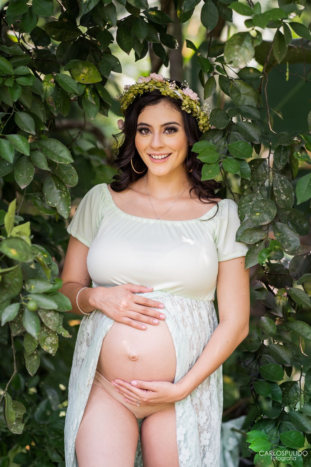 Maternity portrait with floral crown and white lace dress against green garden foliage.