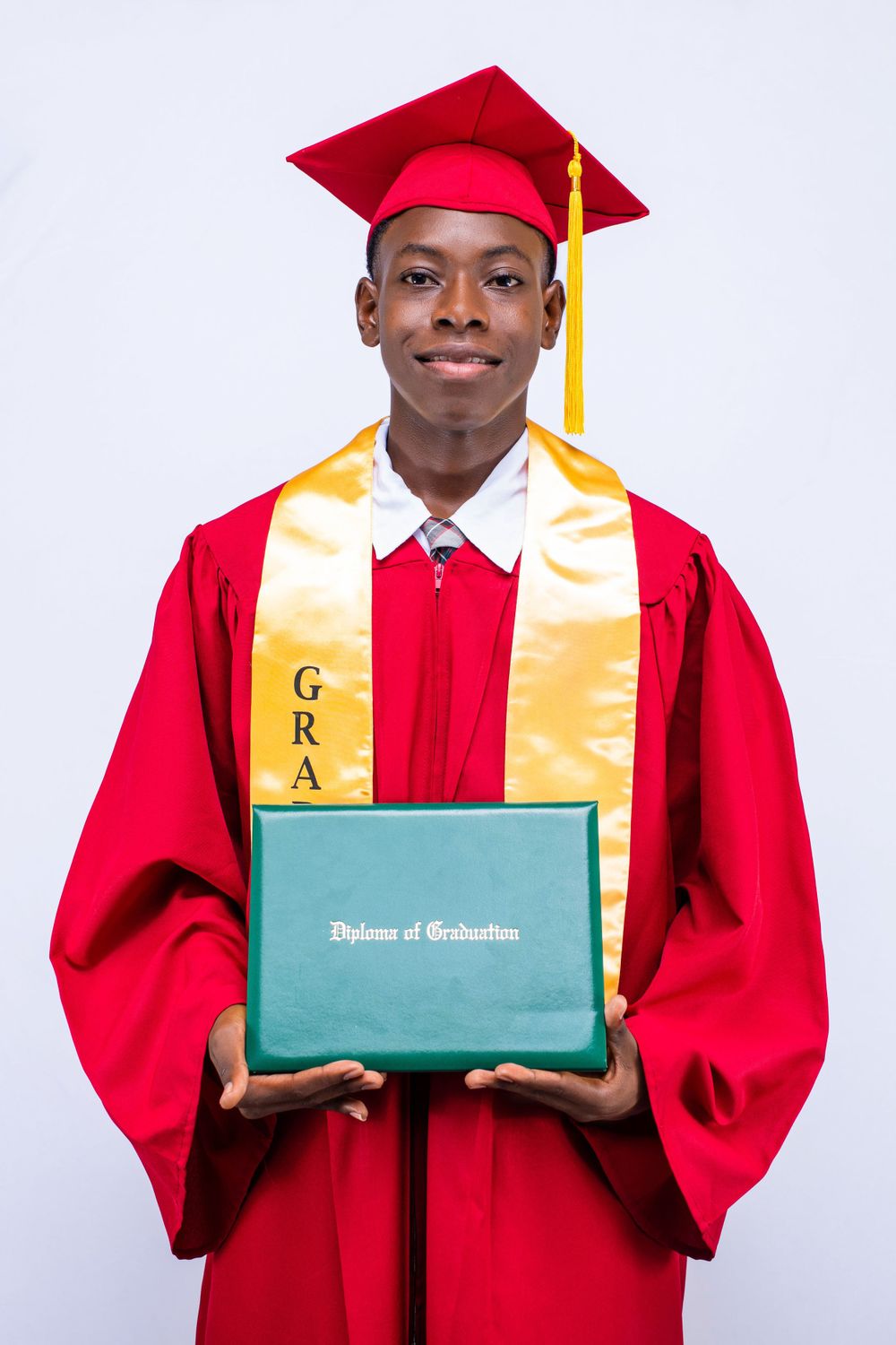 Graduate in red and gold academic regalia holding green diploma cover against white backdrop.