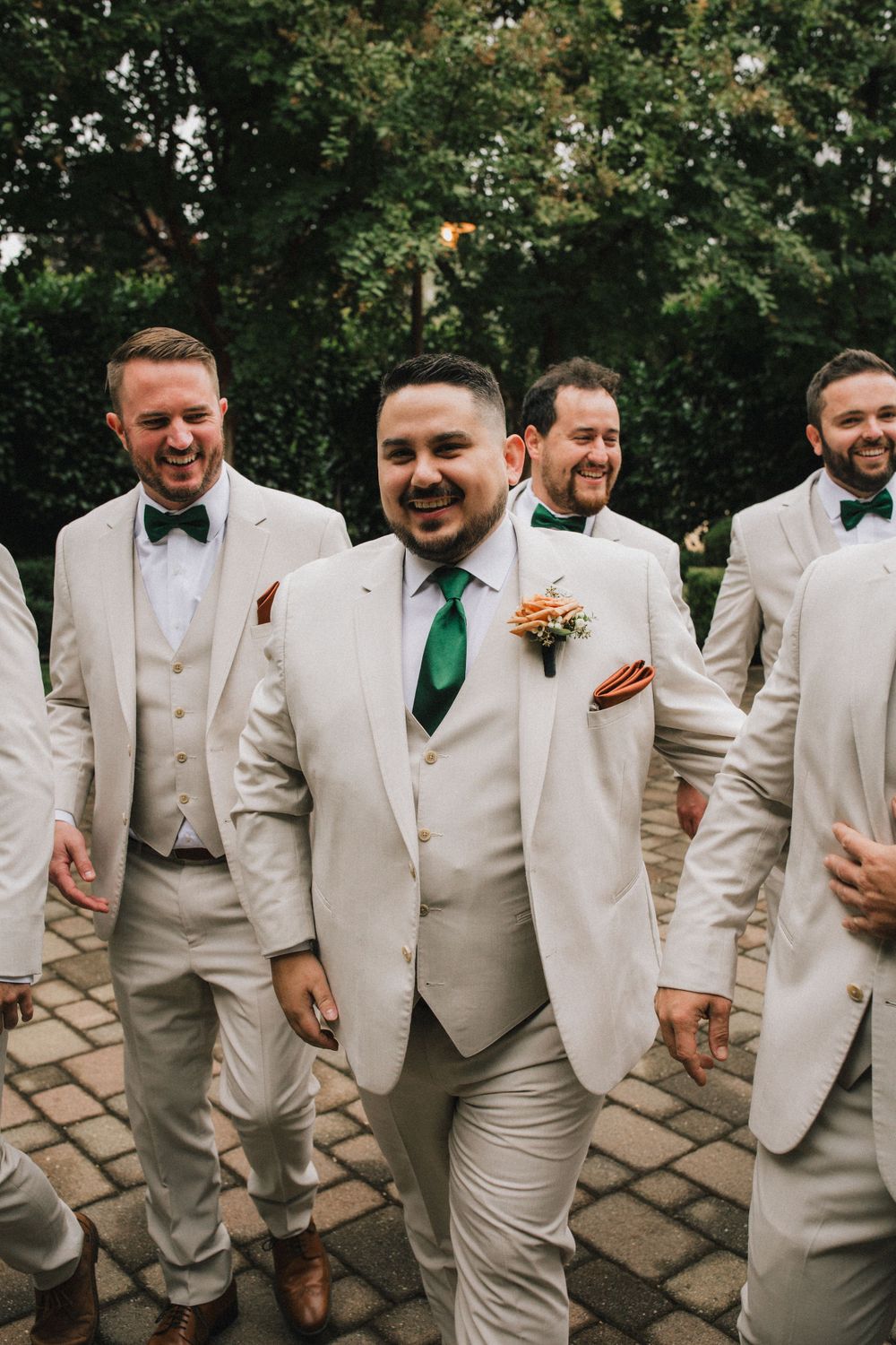 Groomsmen in matching beige suits and bowties share a candid moment on a stone path.