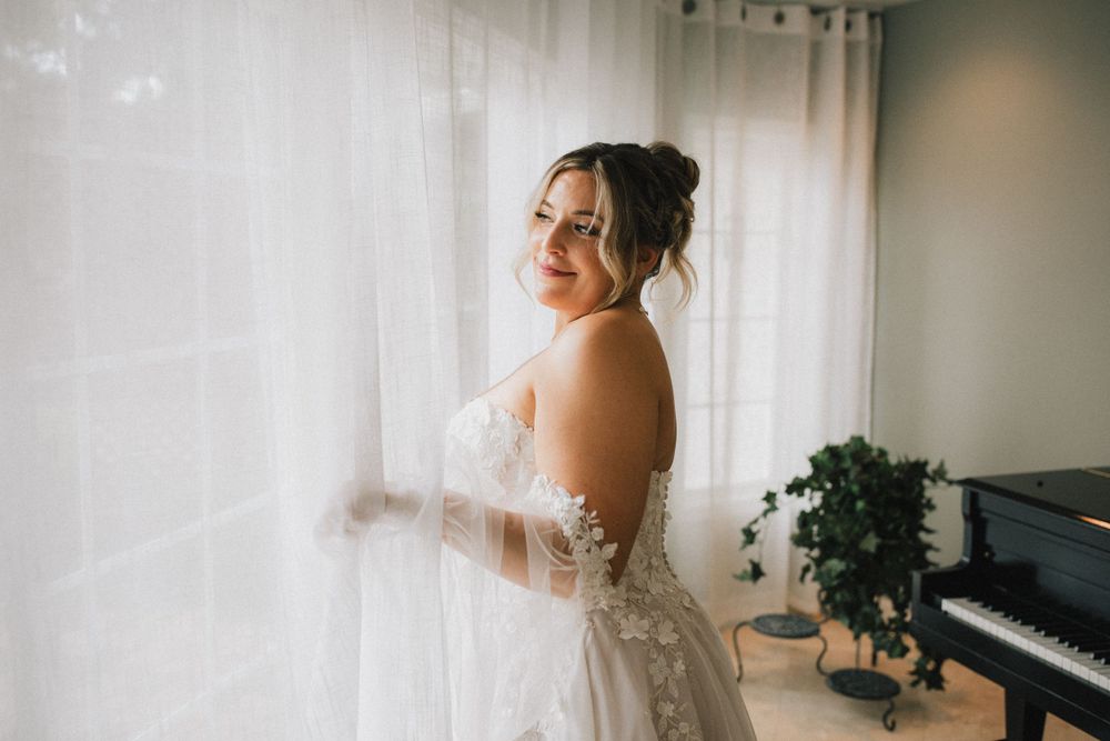 A bride in a lace wedding gown stands by sheer curtains near a piano.