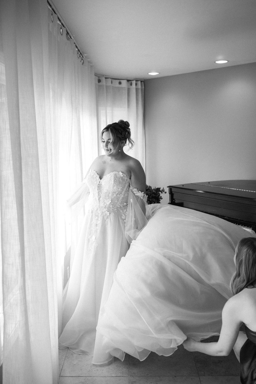 Black and white photo of a bride getting ready in a room with sheer curtains and piano.