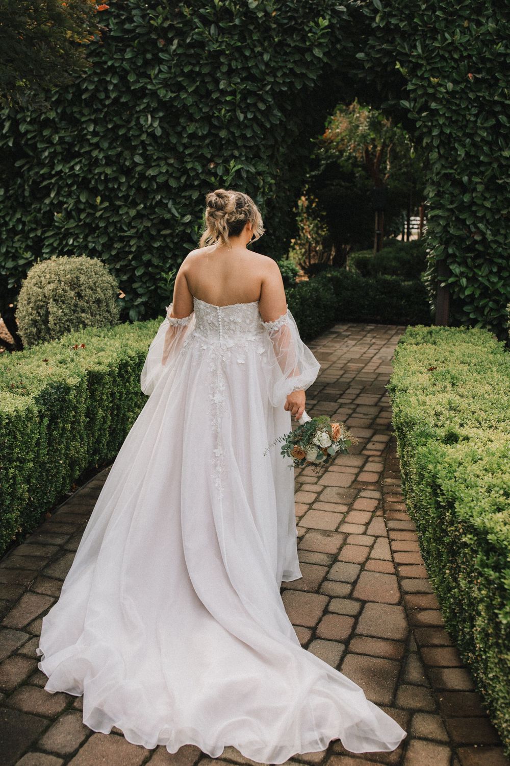 Back view of a bride in flowing white gown walking along stone pathway surrounded by greenery.