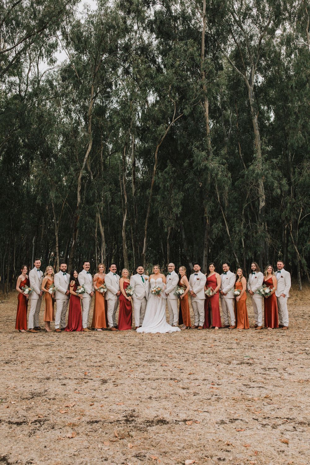 Wedding party in beige and rust colored attire poses in front of tall eucalyptus trees.