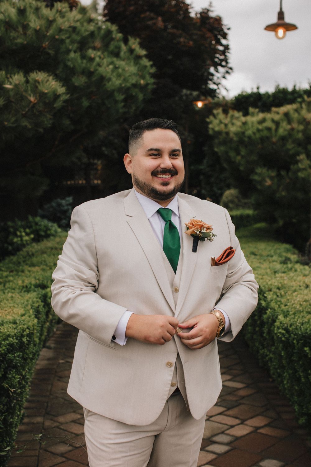 A groom in a light beige suit with emerald green tie smiles while standing outdoors on a stone pathway.