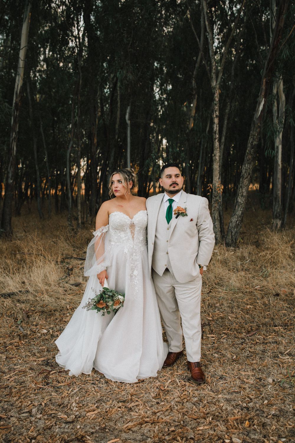 A wedding couple poses elegantly in a eucalyptus forest wearing light-colored formal attire.