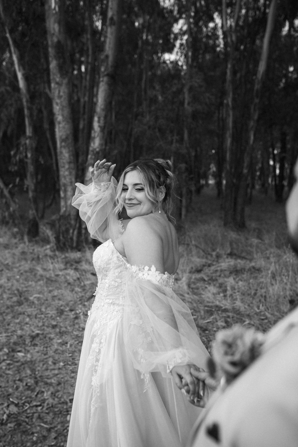 A bride in a flowing white dress smiles joyfully while twirling in a wooded forest setting.