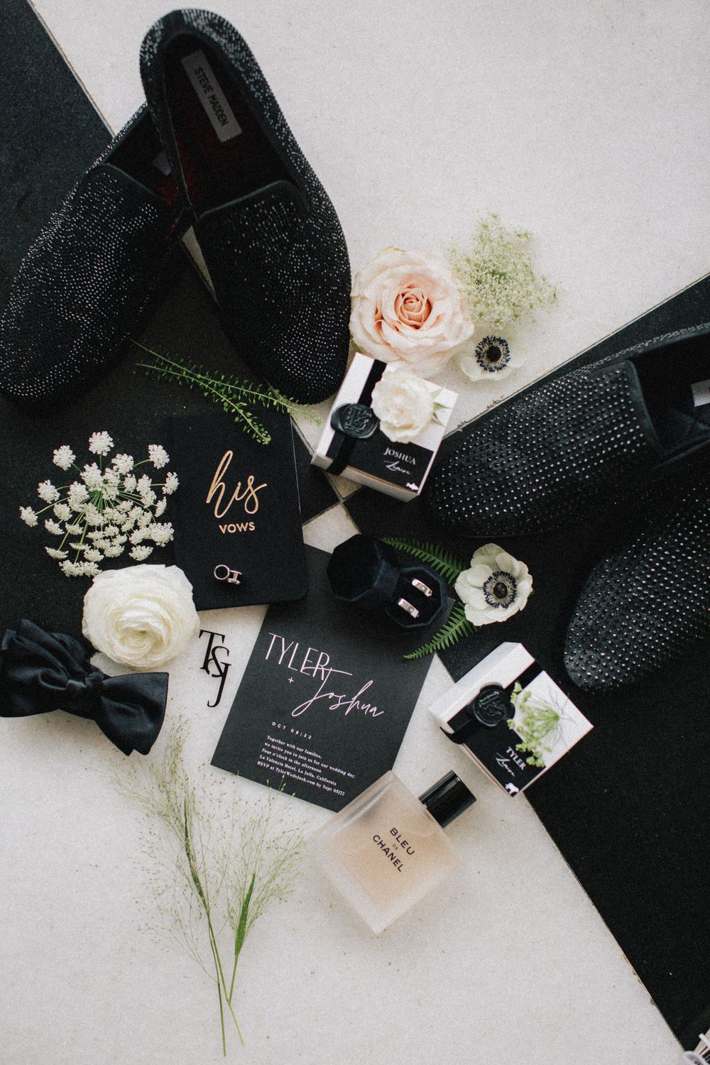 Wedding details flatlay featuring black shoes, invitations, perfume and blush roses on white background.