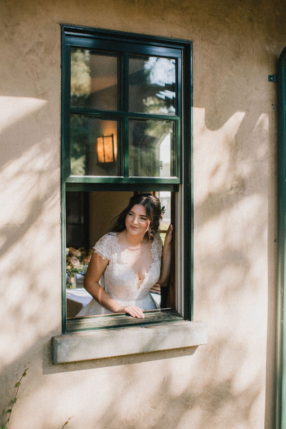 A dreamy bridal portrait framed within a rustic window with dappled sunlight on stucco walls.