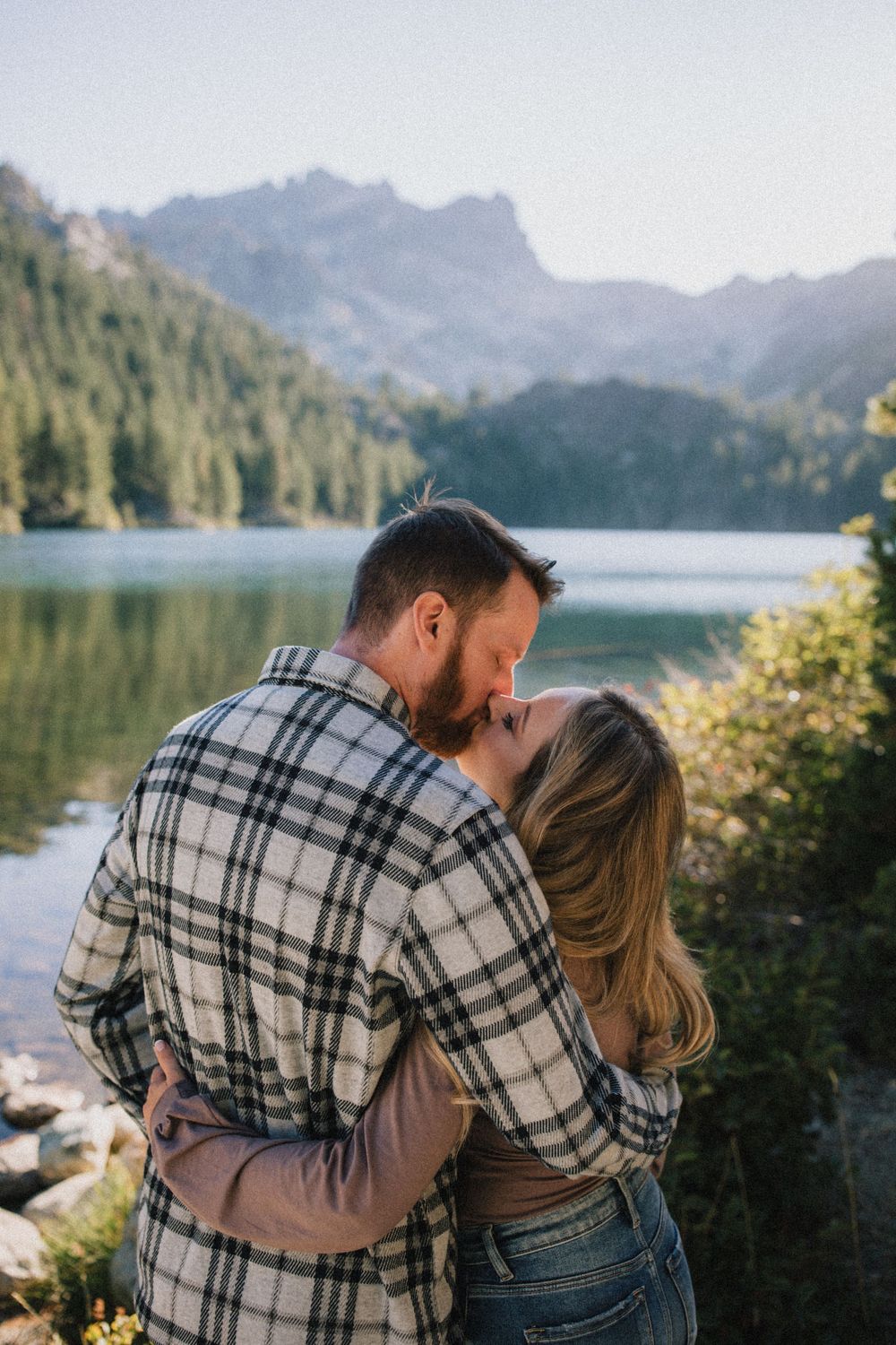 A romantic embrace overlooking a serene mountain lake with evergreen forests in the background.
