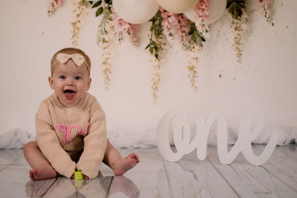 Baby sitting on floor with 'ONE' text decoration and pink balloon backdrop.