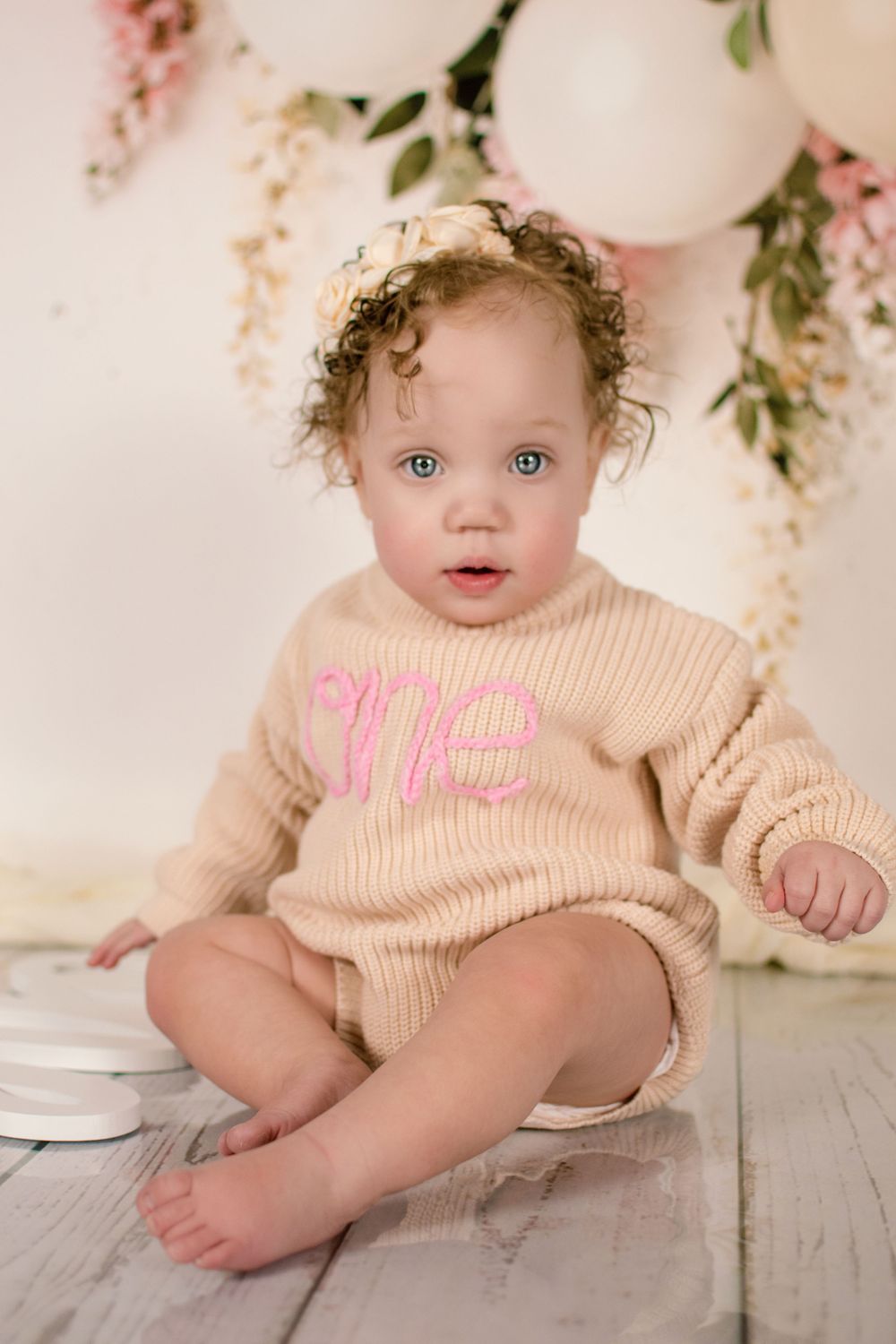 Baby wearing beige outfit sitting on wooden floor with balloon and floral decorations.