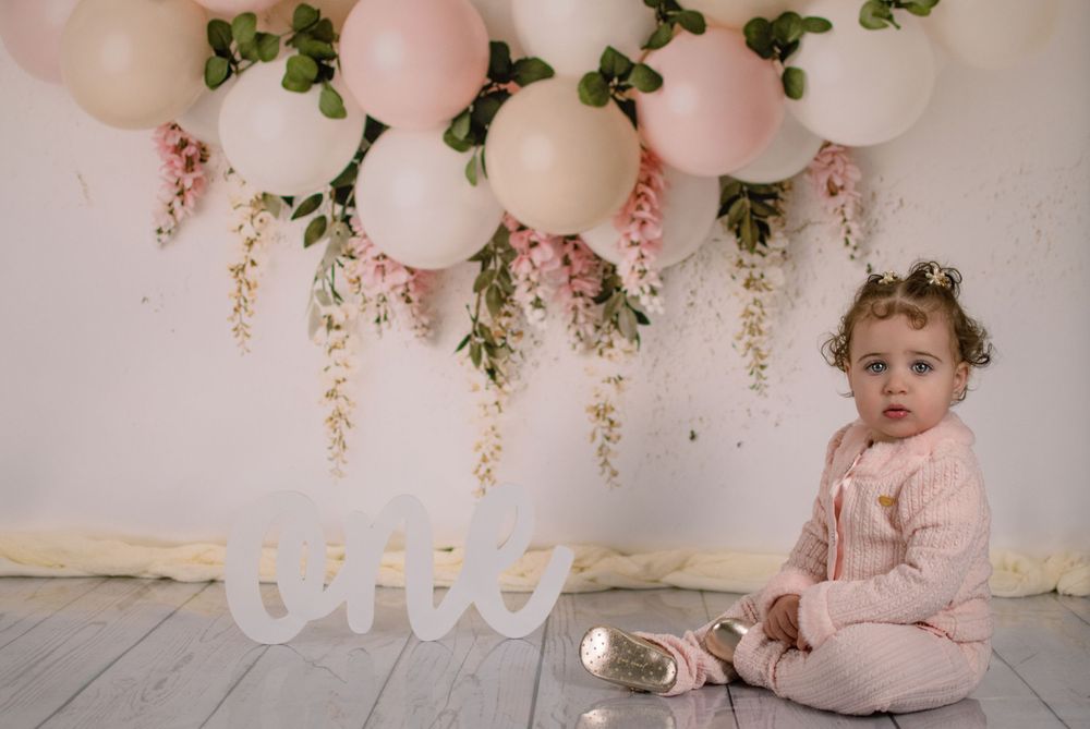 Baby sitting on wooden floor with pink balloons and 'ONE' text decoration.