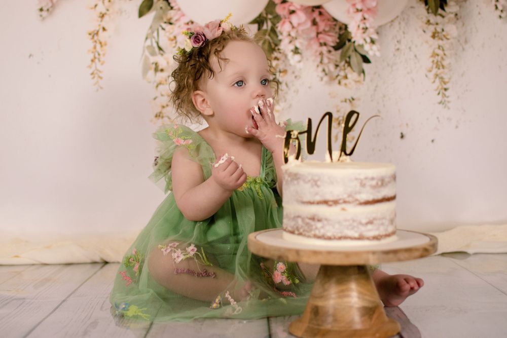 Baby in green dress reaching for birthday cake with balloon and floral decorations.