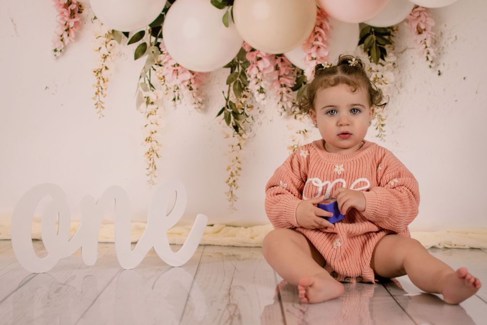 Baby in pink outfit sitting on floor with balloon decorations and 'ONE' text.