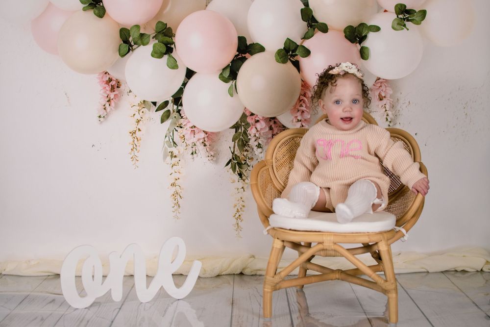 First birthday photo setup with pink balloons, floral decor, and a rattan chair against white backdrop.