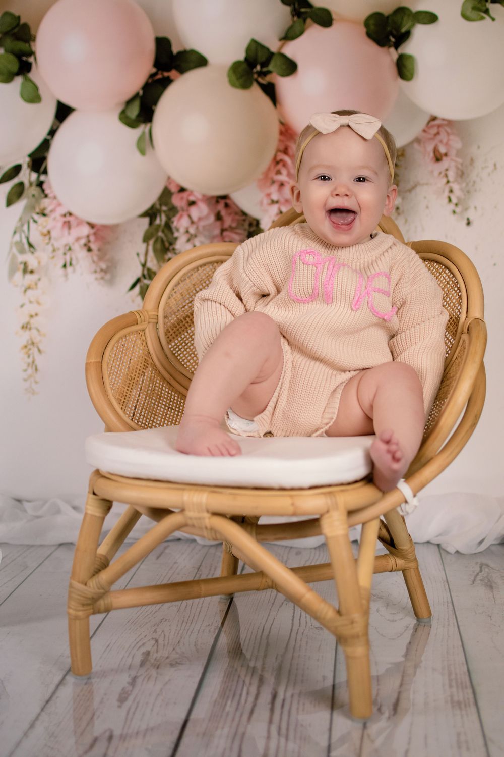 Baby sitting in rattan chair with pink balloon and floral backdrop.