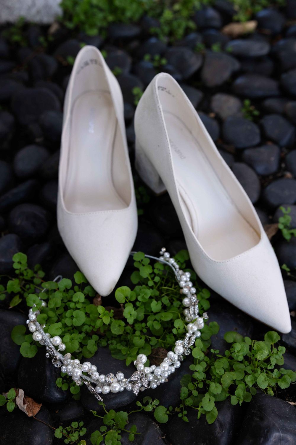 White wedding heels and a delicate pearl hair accessory arranged on black stones with green foliage.