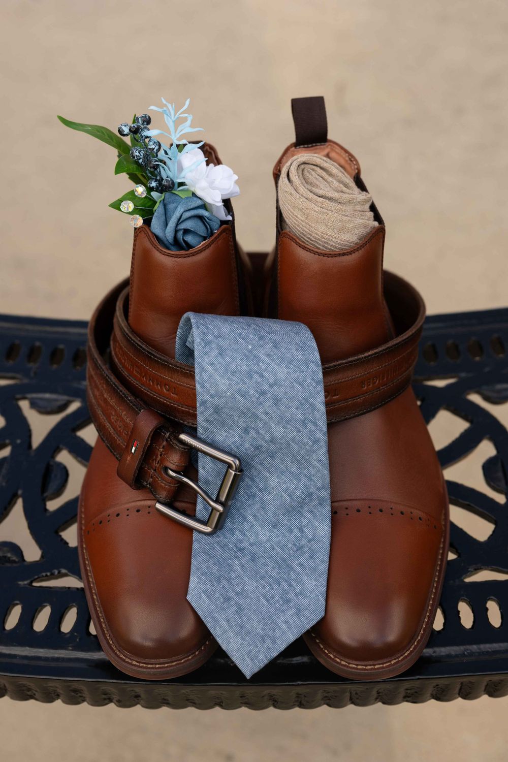 Brown leather dress shoes with a gray tie and blue boutonniere arranged for a wedding.