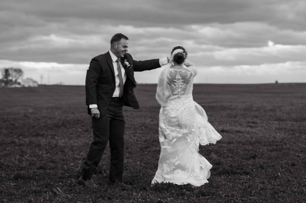 Black and white photo of newlyweds dancing in a windy field under dramatic cloudy skies.